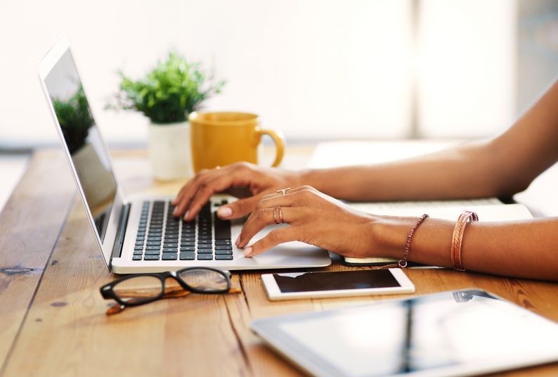 A person's hands rest on a laptop's keyboard. The person, who is out of the frame, is seating at a desk with a plant, mug, phone, glasses and a tablet. 