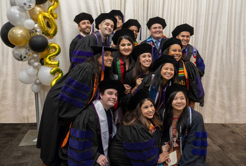 14 graduates wearing regalia pose for a photo in front of a white backdrop 