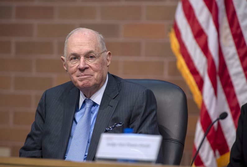 A man in a suit is seated next to an American flag